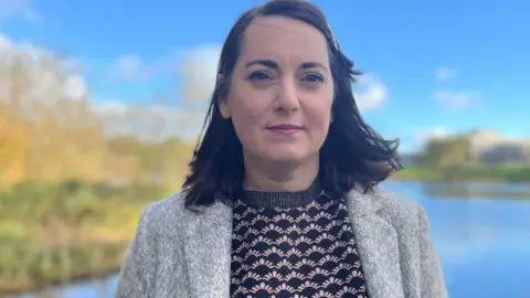 Head and shoulders shot of Prof Niamh Fitzgerald, the report's principal investigator, standing with a serious expressed with a large lake in the background. She is wearing a patterned blouse and a grey cardigan.