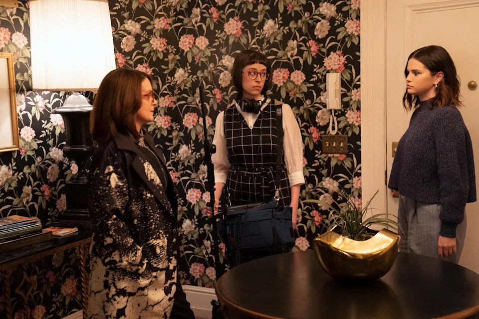 Selena Gomez, an individual in a floral patterned coat, and another person wearing glasses and headphones, stand in a room with flower print wallpaper