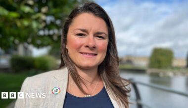 Charlotte Horobin with long brown hair, dark blue top and checked jacket, standing outside in front of the River Nene.