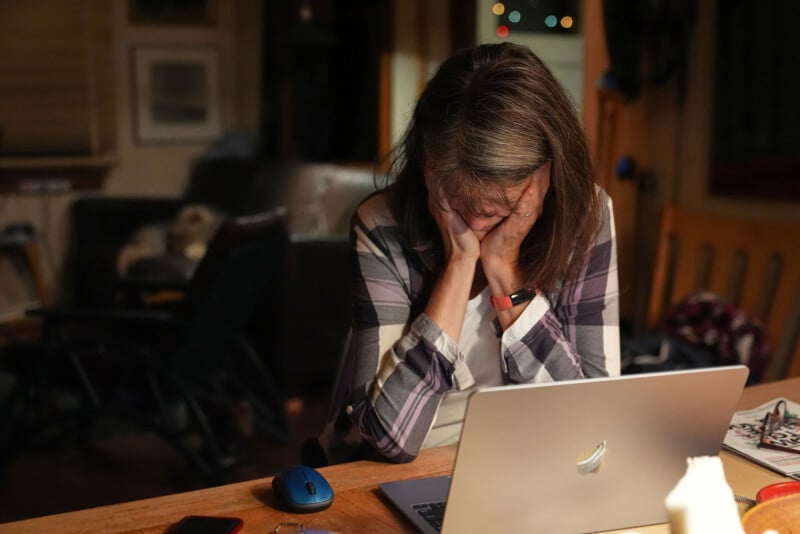 A woman sits at a table with her head in her hands, looking stressed in front of an open laptop. The room is dimly lit and cluttered, suggesting a feeling of overwhelm or frustration.