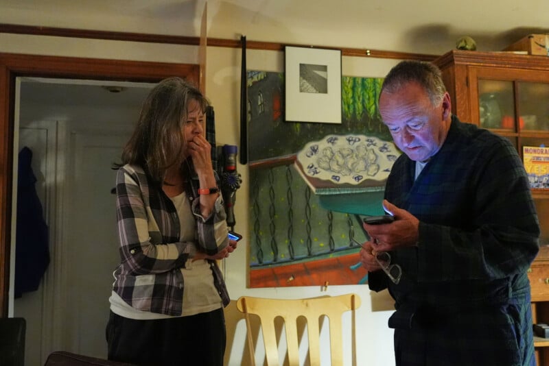A woman stands with her hand on her mouth while a man in a robe looks at a smartphone, both indoors in a warmly lit room with colorful artwork and wooden furniture in the background.