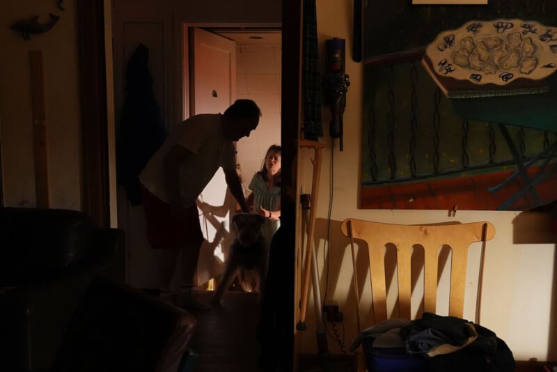A man and a young girl stand in a dimly lit doorway with a dog. The man is reaching toward the dog, and warm light from another room softly illuminates the scene. A painting and wooden chair are visible in the foreground.