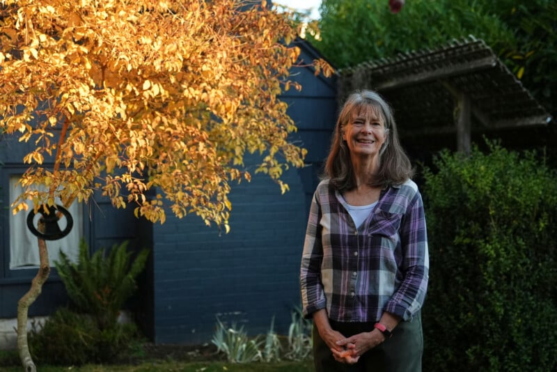 A woman with long gray hair stands smiling in a garden, wearing a plaid shirt over a white top. Behind her is a tree with yellow leaves, a blue wall, and greenery bathed in warm sunlight.