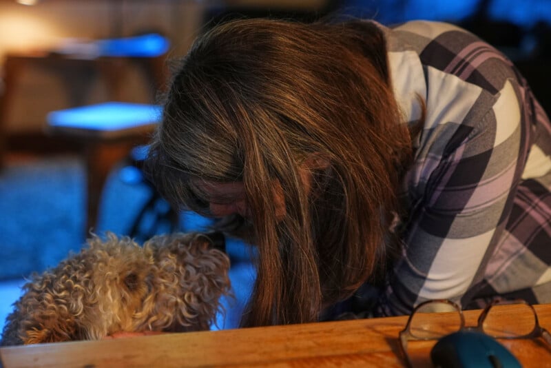 A woman with long brown hair and a plaid shirt gently touches foreheads with a curly-haired dog at a wooden table, creating a tender moment indoors. Glasses and a computer mouse are on the table in the foreground.