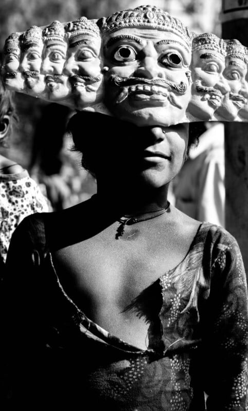 A person wearing a multi-faced traditional mask stands in partial sunlight, casting dramatic shadows across their face and body. The image is in black and white, highlighting the mask’s intricate details.