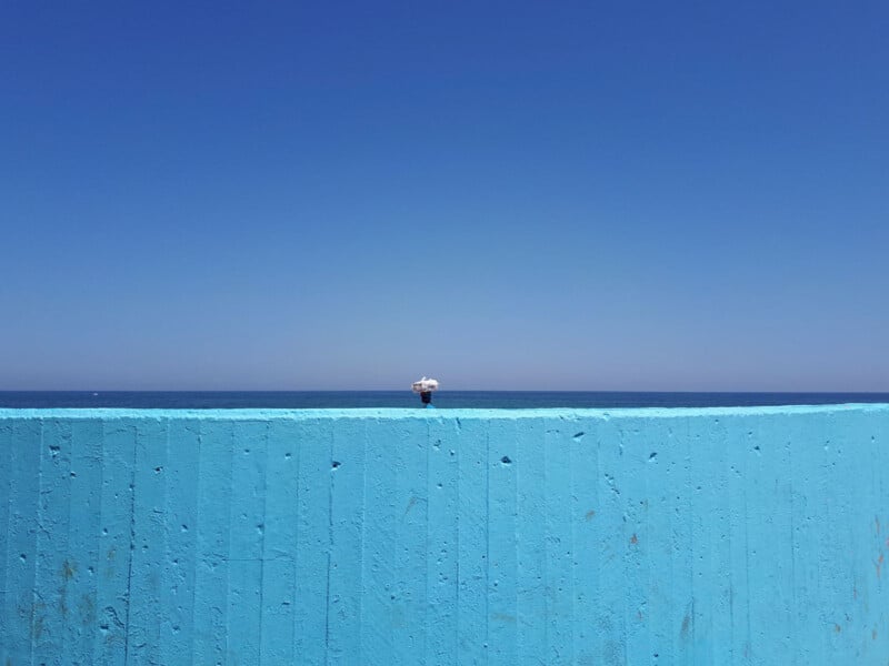 A small white lamp sits on top of a bright blue wall, with the deep blue sea and clear sky in the background under bright sunlight.
