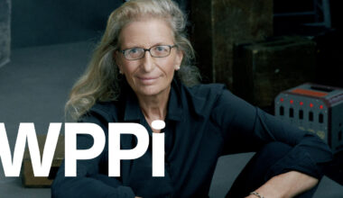 A woman with long, wavy blonde hair and glasses, wearing a black shirt, sits with her arms resting on her knees. Large white letters "WPPI" are overlaid on the image.