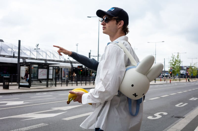A person in sunglasses, a hat, and a white shirt stands by a street pointing ahead, holding bananas and wearing a white Miffy rabbit backpack. Urban background with bus stop and bicycle lane.