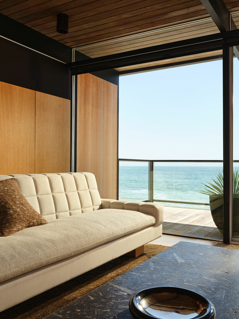 Modern living room with a beige tufted sofa, brown accent pillow, large window with ocean view, wood panel walls, and a potted plant on a balcony.