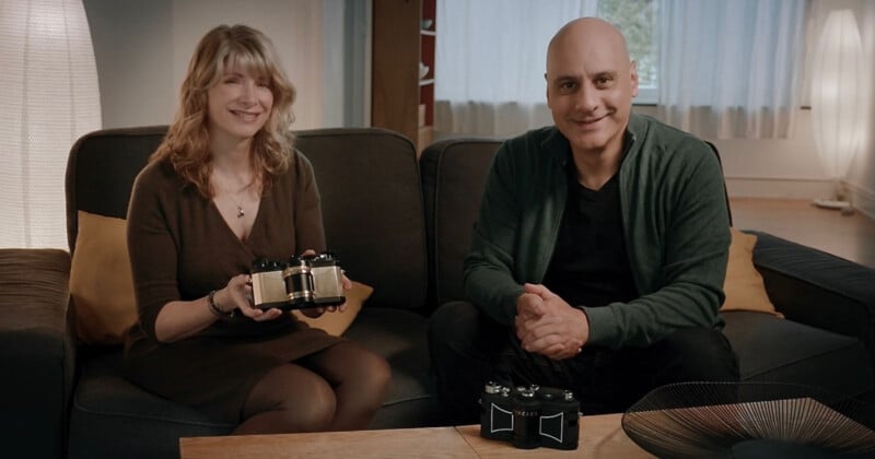 A woman and a man sit on a couch in a cozy living room, smiling at the camera. The woman holds a small robotic device, and another identical device is on the table in front of them.