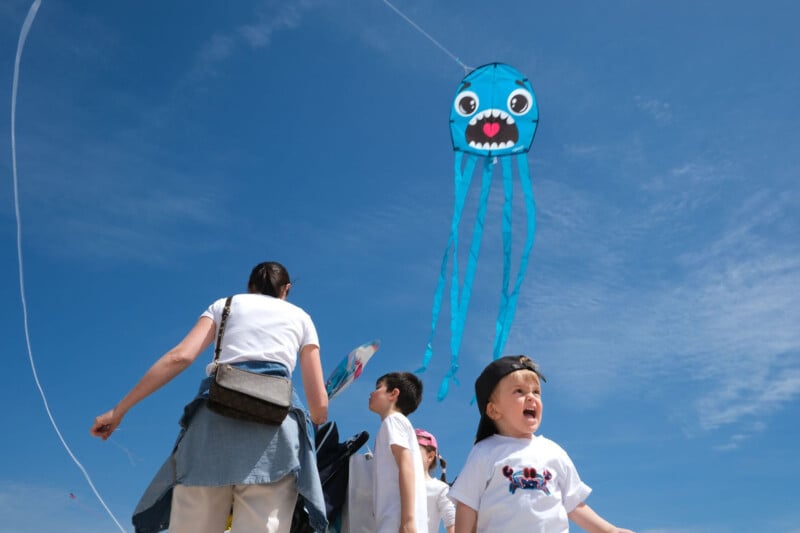 A group of people, including a child in front, fly a blue jellyfish-shaped kite with a cartoon face and long streamers in a clear, sunny sky. The child looks excited, and everyone is dressed casually.