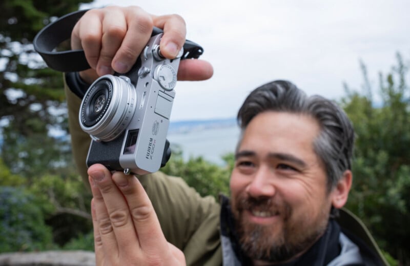 A man with a beard and gray hair smiles while holding a Fujifilm camera outdoors, with greenery and blurred water in the background.