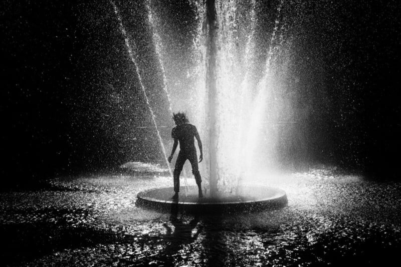 A silhouette of a person stands in the spray of a brightly lit fountain at night, surrounded by mist and water droplets, creating a dramatic, high-contrast scene.