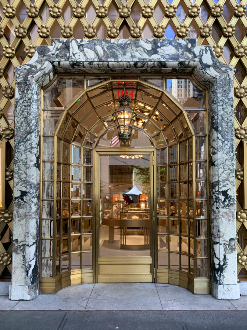 A decorative marble and gold entryway with glass doors and a hanging lantern by Tiffany Woodson leads into a well-lit, upscale interior space.