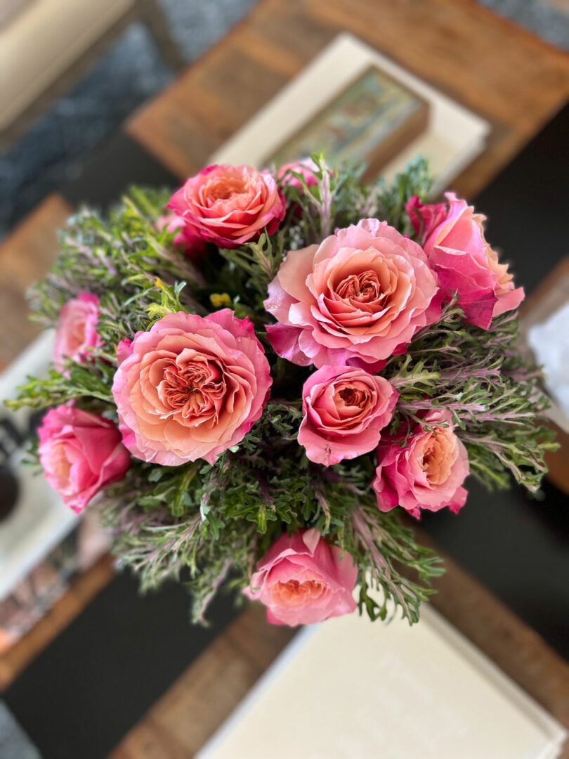 A bouquet of pink and peach roses with green foliage, artfully arranged by Tiffany Woodson, sits elegantly on a wooden table.