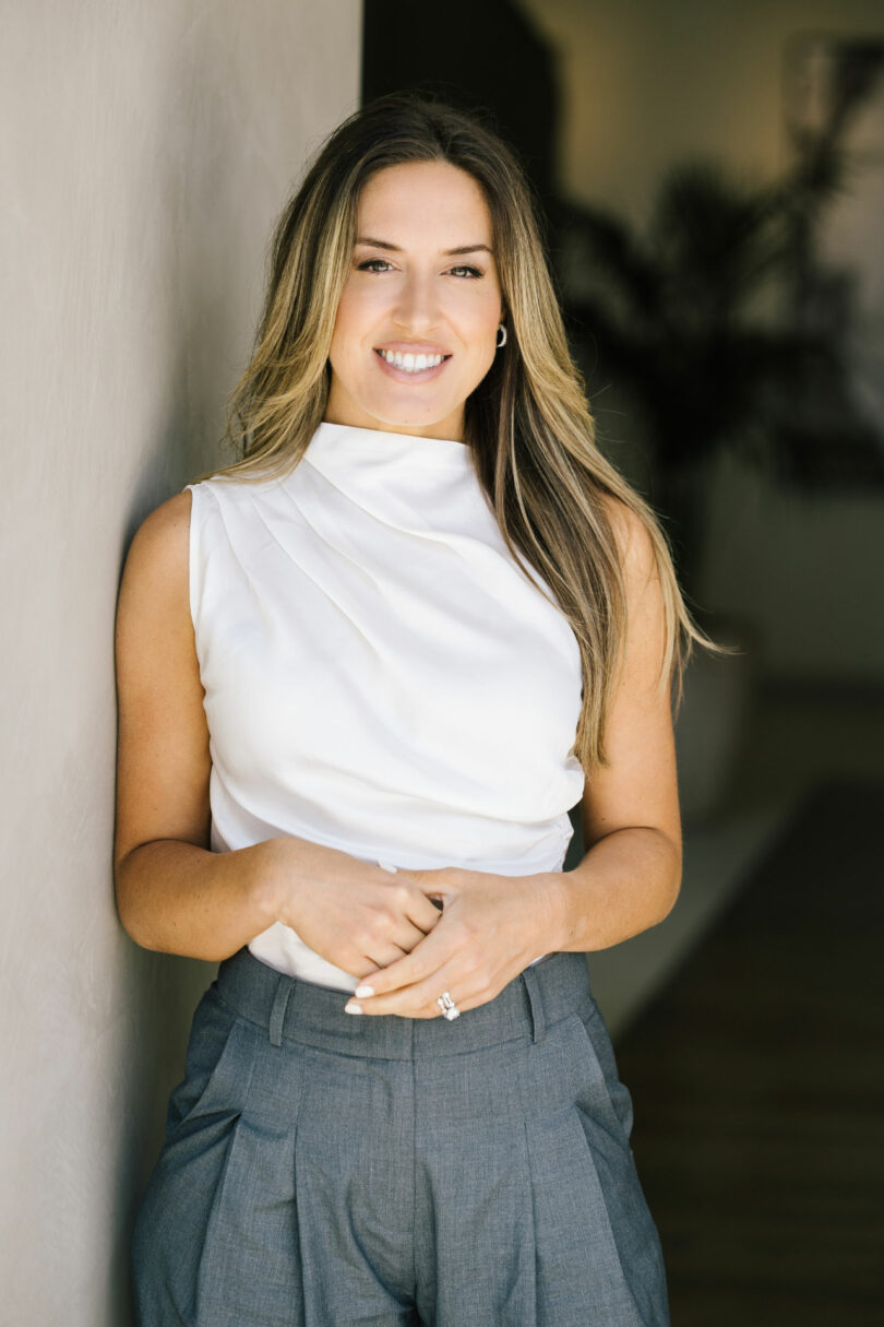 Tiffany Woodson, a woman with long hair wearing a white sleeveless top and gray pants, stands indoors, smiling as she leans against a light-colored wall.