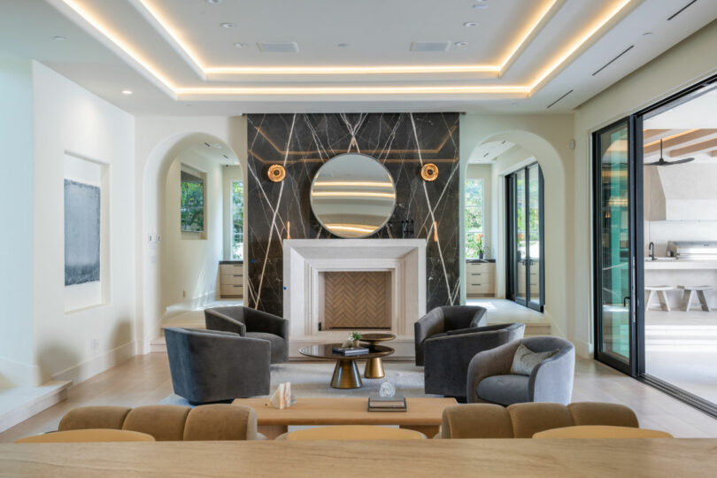 Modern living room designed by Tiffany Woodson, featuring a round mirror above a marble fireplace, gray armchairs, a central coffee table, and large sliding glass doors leading to an inviting outdoor area.