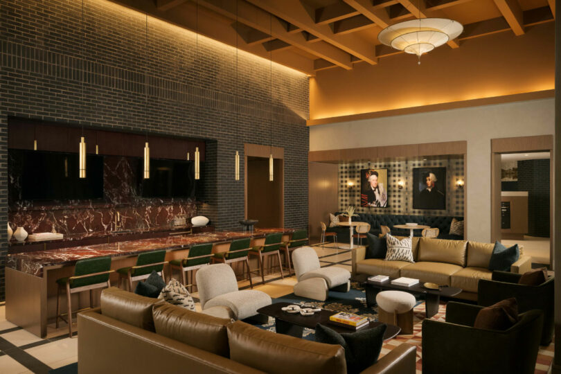 Modern lounge with brown and gold tones, designed by Tiffany Woodson, featuring sofas, armchairs, a marble countertop, dining tables, large TVs, and wall art of two people in suits.