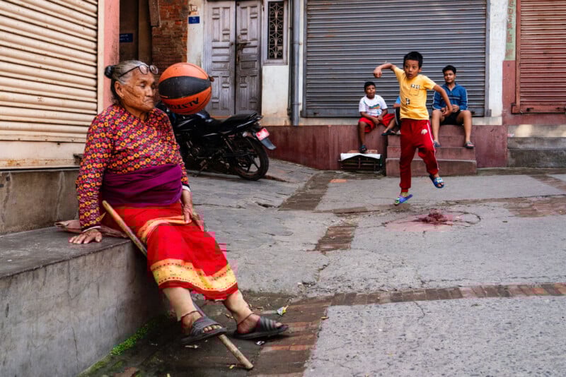 An elderly woman sits on a ledge with closed eyes and a cane, while a boy kicks a ball nearby. Two other boys sit on steps, watching. Shuttered shops and a parked motorcycle are in the background.