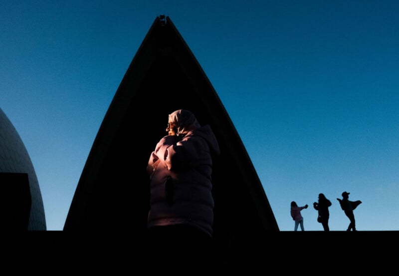 A person in a hooded jacket stands in dramatic light in front of a large, dark architectural structure. Three silhouetted people pose and take photos on the right side against a clear blue sky.