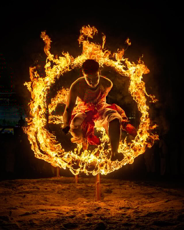 A person in traditional attire is jumping through a large ring of fire at night, with flames dramatically illuminating the scene against a dark background.