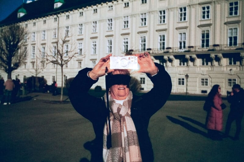 A person in a scarf and dark coat smiles while taking a selfie with a smartphone outside a large, ornate building. Several people stand or walk in the background on a sunny day.