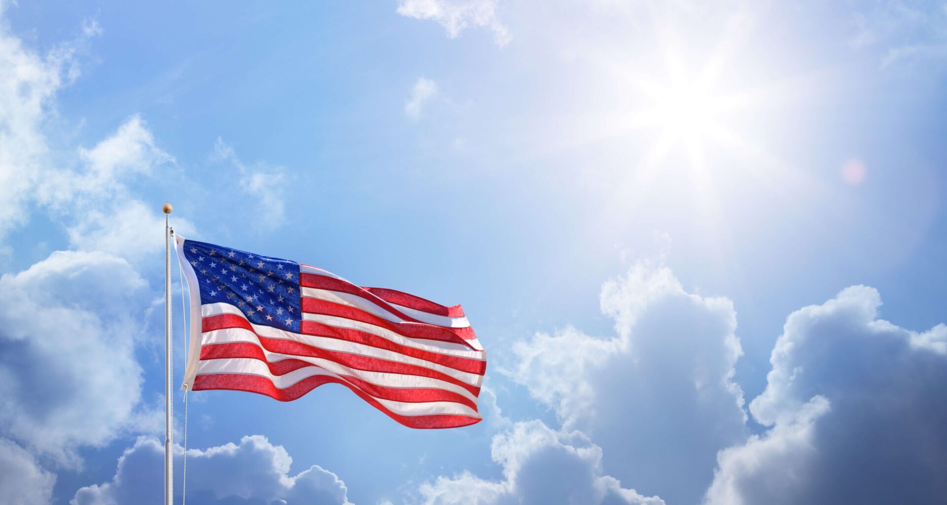 United States flag waving from a flagpole in front of a partially cloudy sky with the sun out