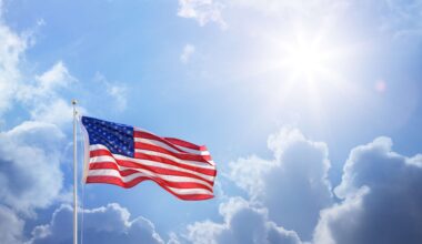United States flag waving from a flagpole in front of a partially cloudy sky with the sun out