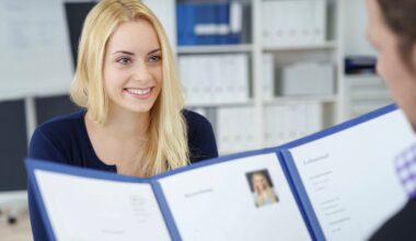 A woman with long blonde hair smiles while sitting across a desk from a person holding a folder with her resume and photo during a job interview in an office setting.