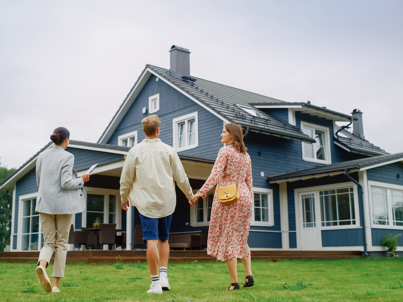 Real Estate Agent Showing a Beautiful Big House to a Young Successful Couple. People Standing Outside on a Warm Day on a Lawn, Talking with Businesswoman, Discussing Buying a New Home.