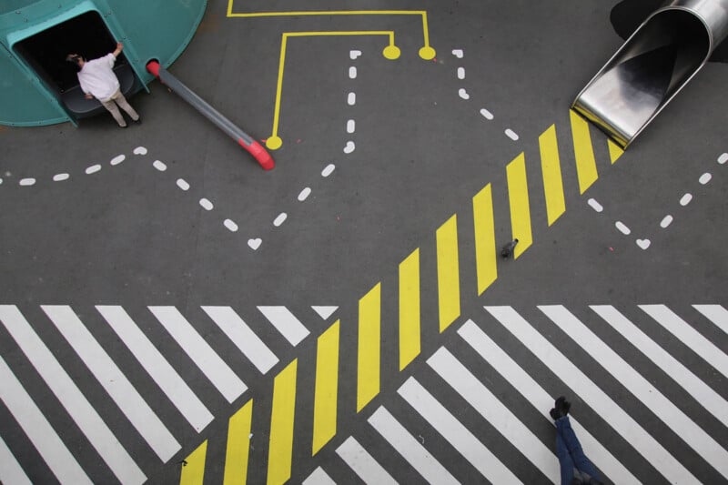 Aerial view of a playground with geometric patterns; yellow and white lines cross the gray ground. A person enters a tunnel on the left, another stands near a slide on the right, and one foot is visible at the bottom.