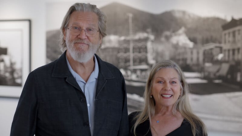 An older man with gray hair and a beard stands next to a smiling woman with long blonde hair. They are indoors, with blurred black-and-white photos and a mountain landscape in the background.