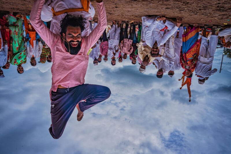 A man jumps energetically in the air with a joyful expression, while a crowd of colorfully dressed people stands behind him outdoors, under a cloudy sky.