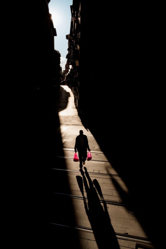 A person carrying two red shopping bags walks along a sunlit street, casting a long shadow, while most of the surroundings are in deep shadow.
