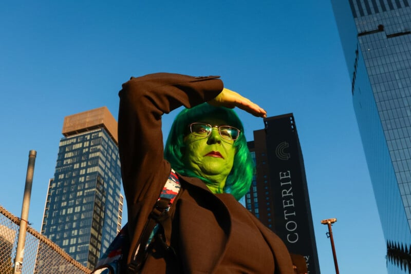 A person with green face paint, turquoise hair, and glasses shields their eyes from the sun while standing in front of tall city buildings under a clear blue sky.