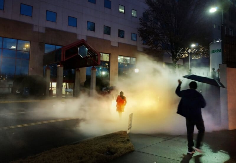 A person in a red jacket walks through thick steam on a city street at dusk, while another person holding an umbrella stands in the foreground near a building with lit windows.