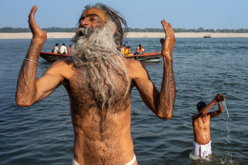 A man with a long gray beard stands waist-deep in a river, eyes closed and arms raised in prayer; another man pours water overhead nearby, with people in a boat and a distant shore in the background.
