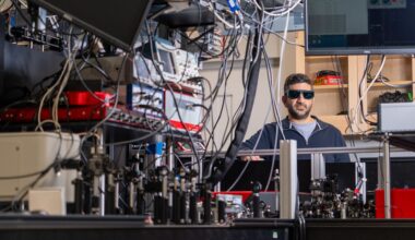 A man wearing dark safety glasses stands behind a table full of wires, mirrors, and lasers