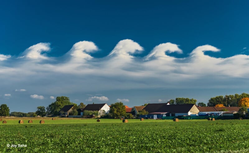 Oddly shaped clouds resembling ocean waves roll across a blue sky above a rural landscape with green fields, hay bales, and houses surrounded by trees.