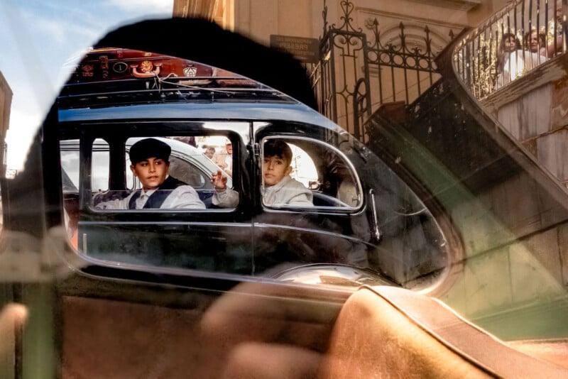 Two boys in vintage clothing sit in the backseat of an old-fashioned car, one giving a peace sign, as seen through a car window with urban reflections and a staircase in the background.