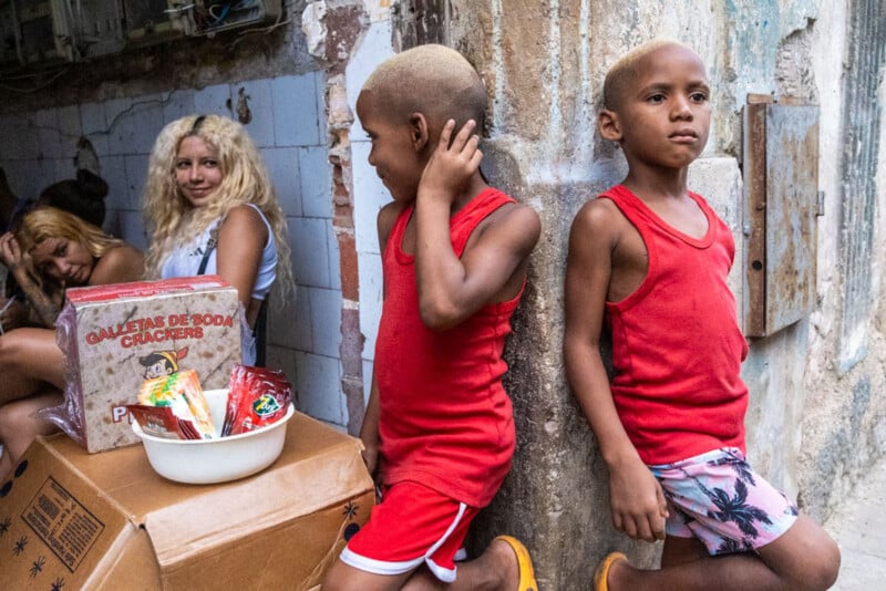 Two young boys in red clothing stand against a weathered wall. Near them, a cardboard box holds snacks and crackers. In the background, two women sit together, one blonde and one with brown hair, inside a tiled, rundown area.