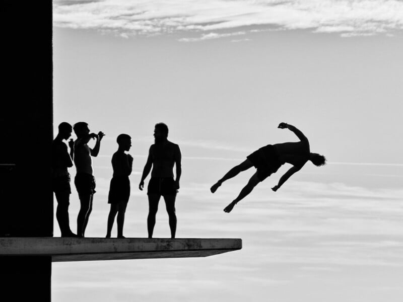A group of five people stands on a diving platform as one person jumps off, captured mid-air against a cloudy sky in black and white.