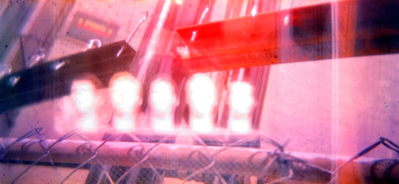 A blurry, abstract photo with five white mannequin heads behind a chain-link fence, vivid light leaks, and distorted red and purple tones. The background contains metal beams and a brick wall.