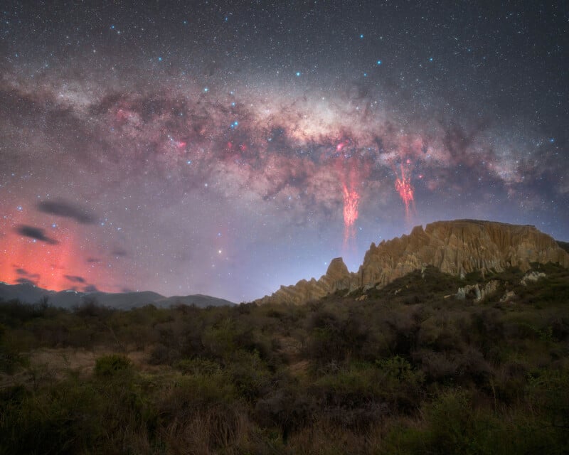 A starry night sky with the bright arc of the Milky Way stretches above rocky hills and desert vegetation; red lightning-like streaks, known as sprites, reach down from the stars in the background.
