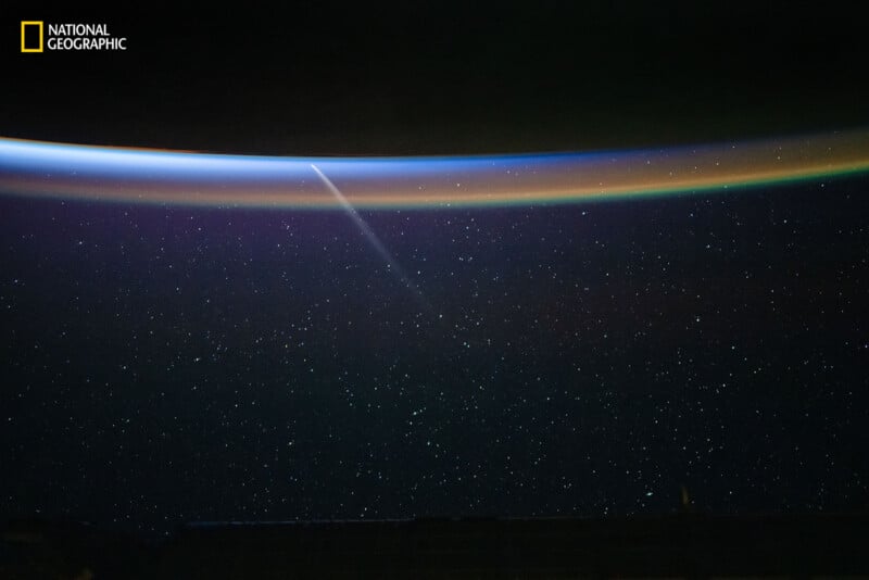 A star-filled night sky viewed from space, with a bright streak cutting through a thin, colorful atmospheric horizon. The National Geographic logo appears in the upper left corner.