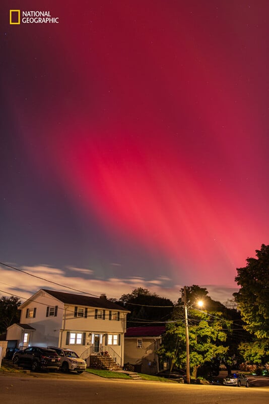 A suburban street at night with a house in the foreground and dramatic red and pink aurora lights illuminating the sky above. The National Geographic logo appears in the upper left corner.