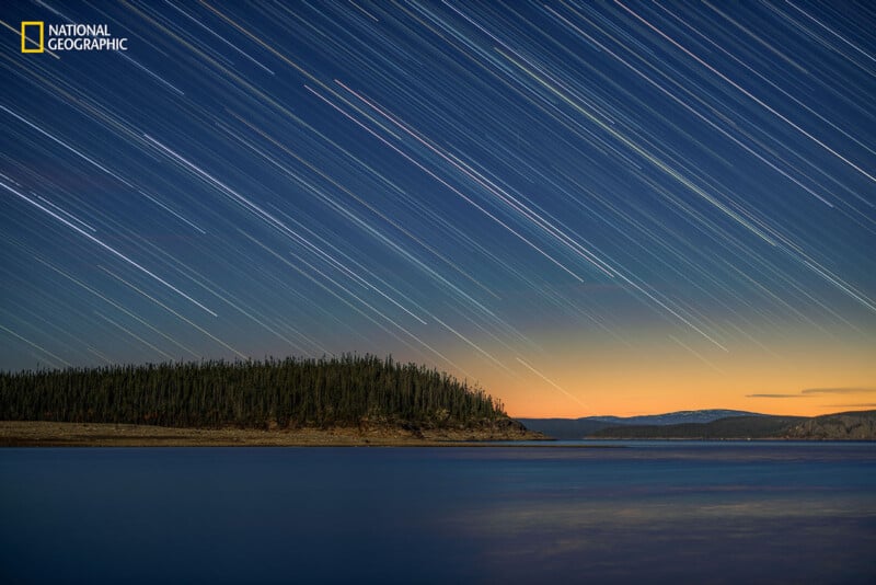 Long-exposure photo of stars leaving light trails over a calm lake, with a forested shoreline and a soft orange glow on the horizon at dusk. National Geographic logo is in the top left corner.