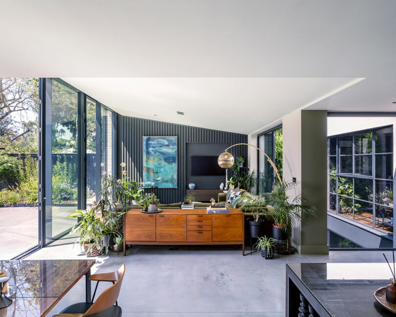 Modern living room with large glass windows, mid-century wooden sideboard, indoor plants, armchair, and wall-mounted TV. Natural light fills the space, highlighting greenery outside.