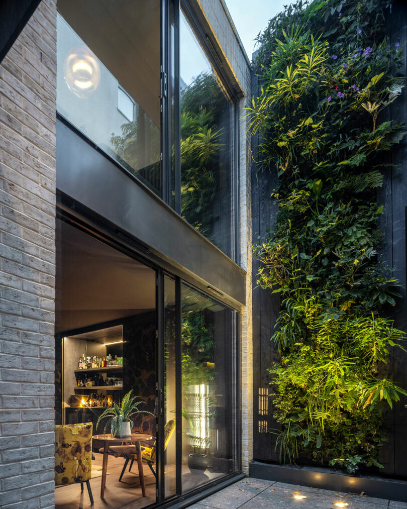 A modern home with large glass doors opens to a patio featuring a vertical garden wall filled with various green plants and accent lighting.