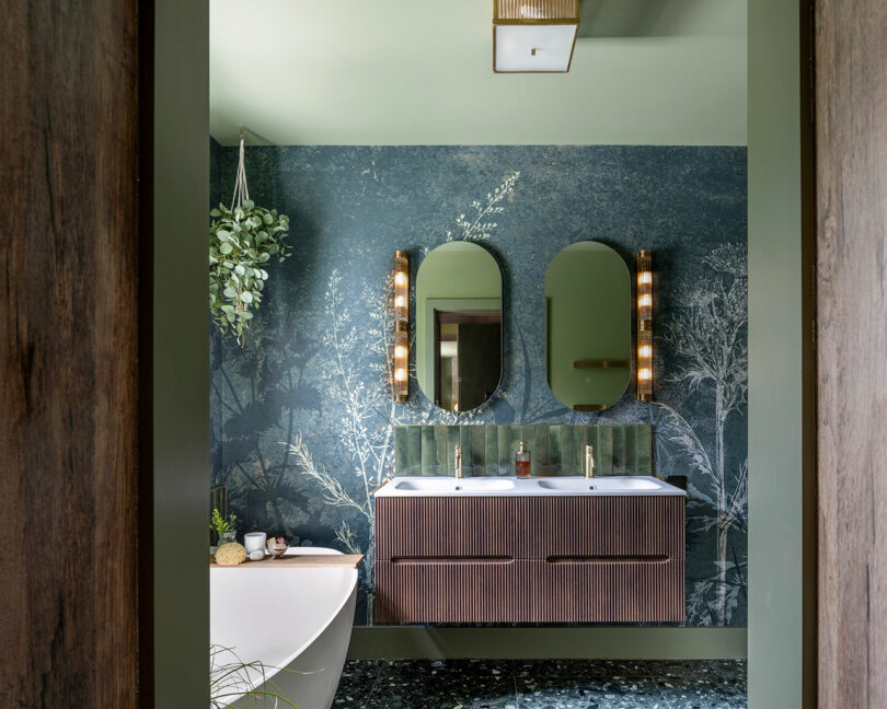 Modern bathroom with a double vanity, two oval mirrors, wall sconces, a botanical mural, a hanging plant, and part of a freestanding bathtub visible in the foreground.
